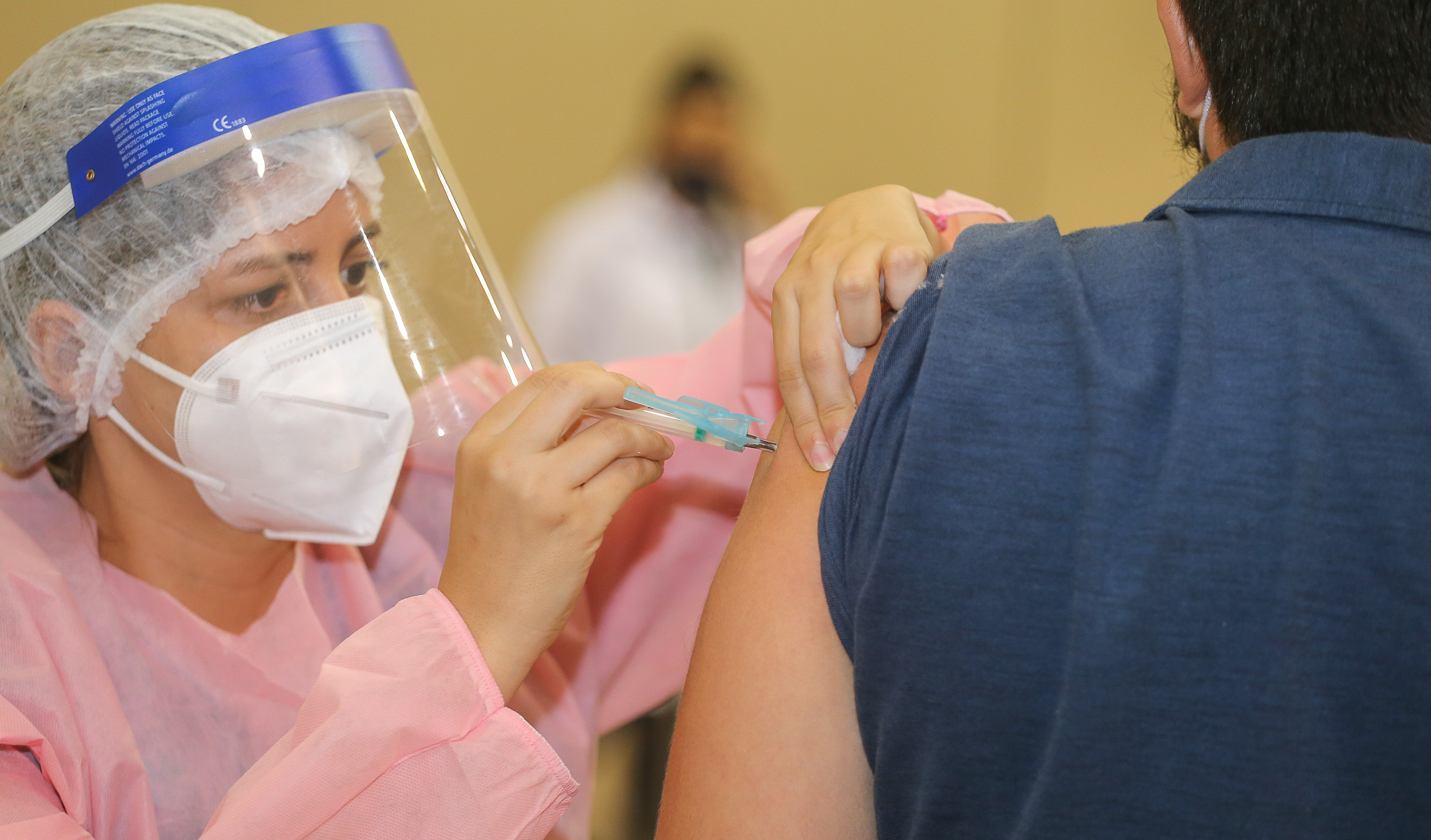 enfermeira de jaleco, máscara, touca e faceshield aplicando vacina em braço de homem de costas
